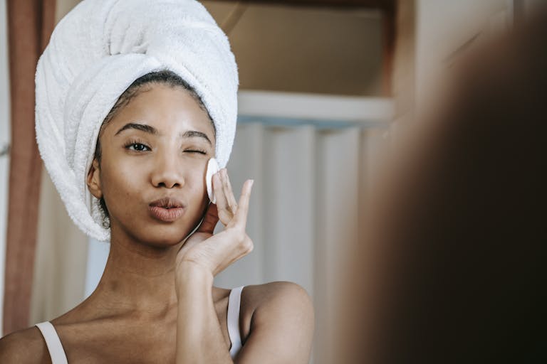 charming-ethnic-female-with-folded-lips-winking-while-cleaning-face-with-cotton-pad-during-daily-beauty-routine
